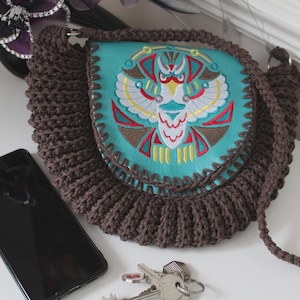 May include: A brown crocheted shoulder bag with a turquoise flap featuring an embroidered owl design in red, yellow, and brown. A black smartphone and keys are next to the bag. The bag has a long, crocheted strap.