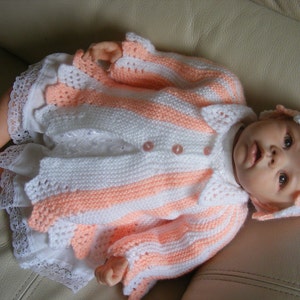 May include: A knitted baby outfit in pink and white stripes with a matching hat. The outfit has a crocheted lace trim and buttons.