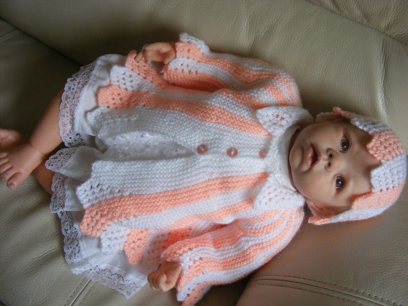 May include: A knitted baby outfit in pink and white stripes with a matching hat. The outfit has a crocheted lace trim and buttons.