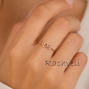 May include: A delicate rose gold ring featuring a musical note design with a small, round, red gemstone. The ring is worn on a finger, showcasing its elegant and minimalist style. The brand name "Rachveli" is written on the hand.