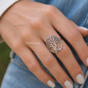 Peut inclure: Bague argentée ornée d'un motif d'arbre de vie ovale. La bague est portée sur une main avec du vernis à ongles blanc, sur un fond en jean. La marque "Rachveli" est visible.