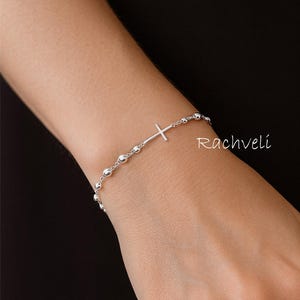 Pode incluir: Uma pulseira de prata com um pingente de cruz e pequenas contas redondas. A pulseira é exibida em um pulso contra um fundo escuro. A marca "Rachveli" é visível.