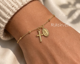 Puede incluir: Pulsera dorada con un dije de cruz y un medallón con una figura religiosa. La pulsera tiene una cadena delicada con pequeñas cuentas redondas. El nombre de la marca "Rachveli" es visible.