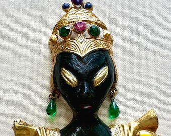 May include: A gold-toned brooch depicting a stylized black woman wearing a crown and earrings with colorful gemstones. The woman's face is painted black with gold accents and the brooch has a ruffled collar with colorful gemstones.
