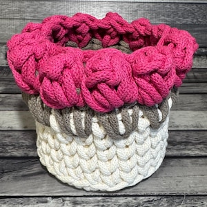 May include: A handmade, round crochet basket in shades of white, gray, and hot pink. The top edge features a decorative, knotted design in bright pink. The basket's body is a textured, woven pattern.