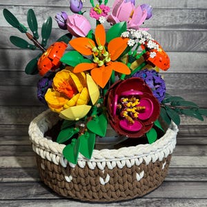 May include: A colorful bouquet of flowers made from plastic building blocks, arranged in a brown and white woven basket. The flowers include orange, pink, yellow, and purple blooms with green leaves. The basket has a white rim and a brown body with white decorative stitching.
