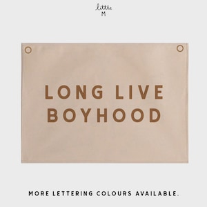 Op de afbeelding: Een beige canvas wandtapijt met bruine tekst die "LONG LIVE BOYHOOD" luidt. Het tapijt heeft twee metalen ringen aan de bovenkant om op te hangen.