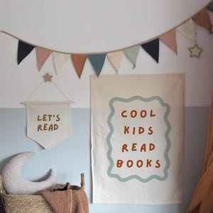 Puede incluir: Una escena de habitaci&oacute;n infantil con una pancarta que dice "LET'S READ" y un p&oacute;ster m&aacute;s grande que dice "COOL KIDS READ BOOKS". Una cesta tejida contiene una almohada de media luna y una manta. Una guirnalda de banderines de colores cuelga arriba.
