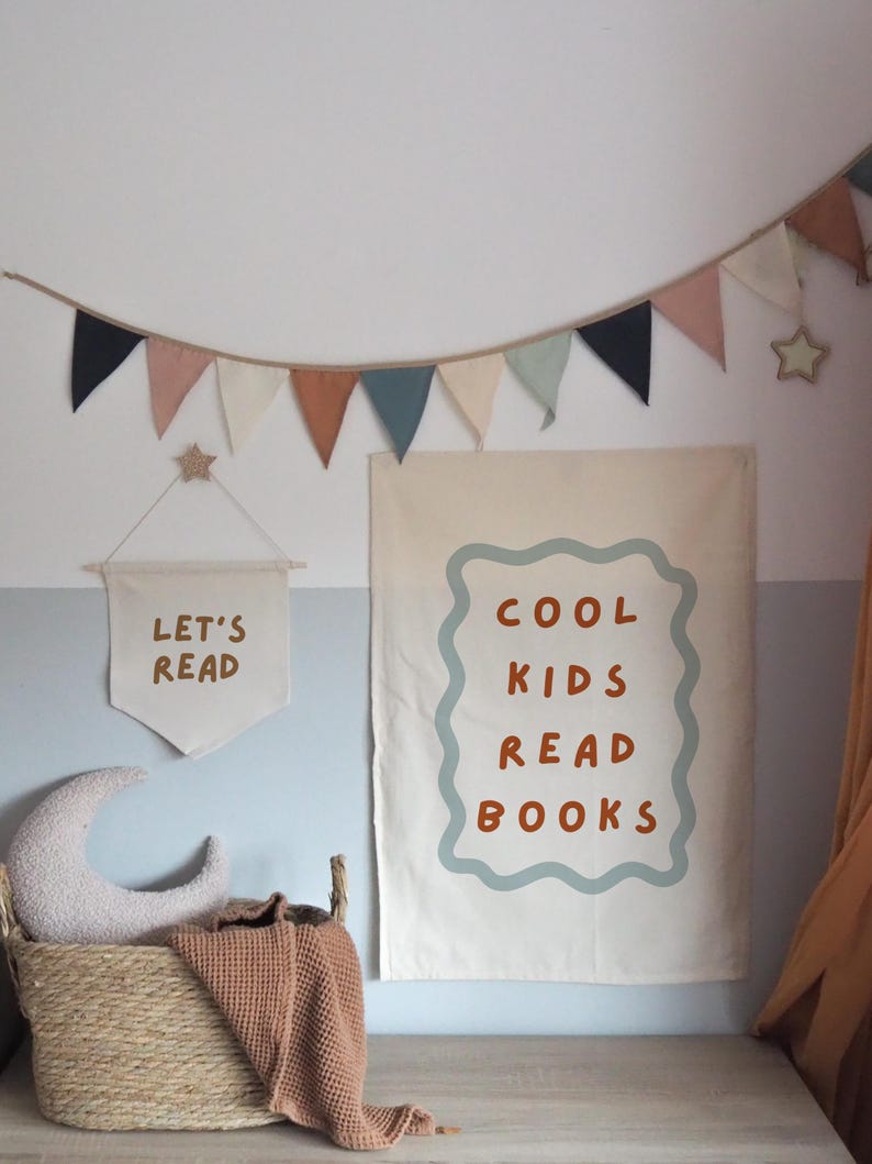 Puede incluir: Una escena de habitaci&oacute;n infantil con una pancarta que dice "LET'S READ" y un p&oacute;ster m&aacute;s grande que dice "COOL KIDS READ BOOKS". Una cesta tejida contiene una almohada de media luna y una manta. Una guirnalda de banderines de colores cuelga arriba.
