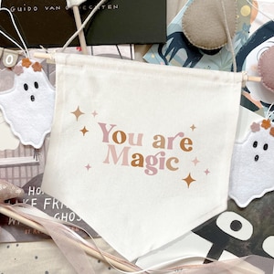 Puede incluir: Una pancarta de lona blanca con las palabras "You are Magic" en letras rosas y marrones, acentuada con formas de estrellas. La pancarta está sujeta por clavijas de madera. Fantasmas decorativos de fieltro y otros elementos artesanales rodean la pancarta.
