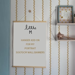 Puede incluir: Un cartel de pared con el texto "little M" y "HANGER ADD ON FOR MY PORTRAIT 50x70CM WALL BANNERS". El cartel est&aacute; suspendido por un colgador de madera. El fondo presenta una pared blanca con rayas onduladas doradas.