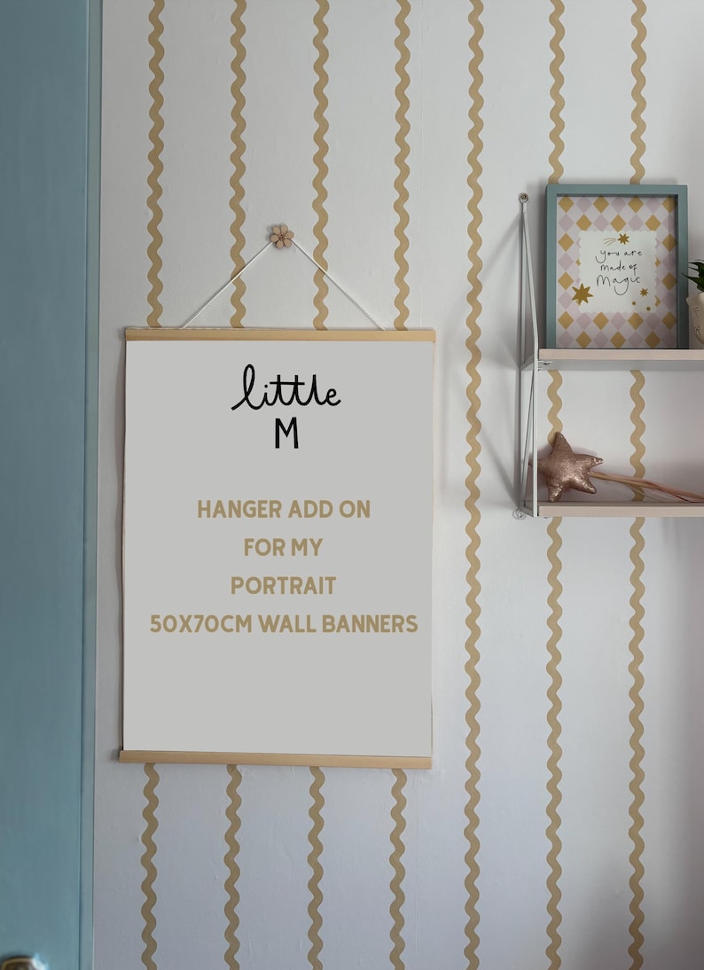 Puede incluir: Un cartel de pared con el texto "little M" y "HANGER ADD ON FOR MY PORTRAIT 50x70CM WALL BANNERS". El cartel est&aacute; suspendido por un colgador de madera. El fondo presenta una pared blanca con rayas onduladas doradas.