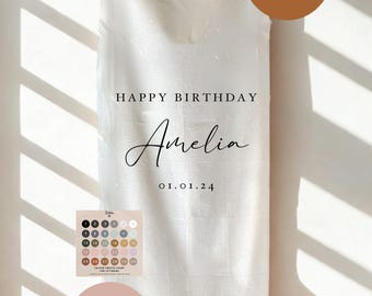 May include: A white fabric banner with the text "HAPPY BIRTHDAY Amelia 01.01.24" in black script. The banner includes color charts and options for lettering. Two brown and pink circles with text are also visible.