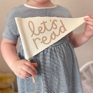 May include: A child holds a white pennant with the words "let's read" in brown lettering. The pennant is attached to a wooden stick.