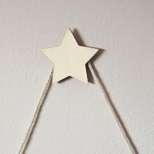 May include: A wooden star with a white finish is hanging from two white strings against a white wall.