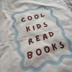 Puede incluir: Bolsa de lona color crema con el texto "COOL KIDS READ BOOKS" en letras granates dentro de un borde ondulado azul claro. La bolsa tiene un aspecto ligeramente arrugado.