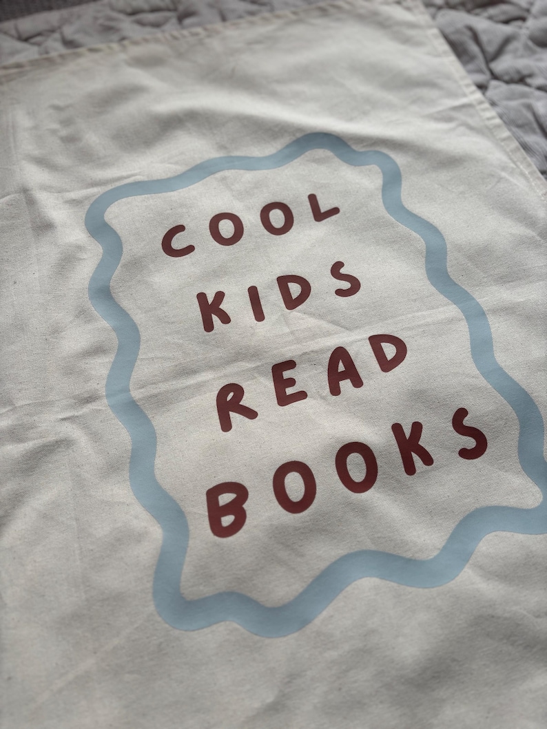 Puede incluir: Bolsa de lona color crema con el texto "COOL KIDS READ BOOKS" en letras granates dentro de un borde ondulado azul claro. La bolsa tiene un aspecto ligeramente arrugado.
