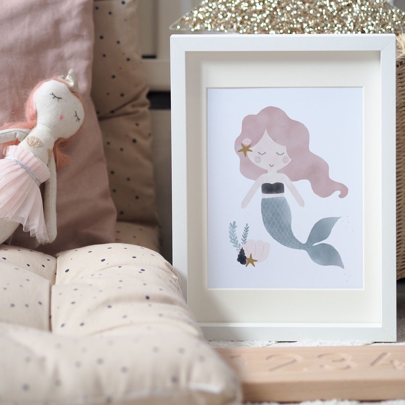 May include: A white framed print featuring a watercolour illustration of a mermaid with pink hair and a teal tail. The mermaid is surrounded by seashells and starfish.