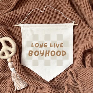 Puede incluir: Una pancarta de lona blanca con un patrón a cuadros y el texto "LONG LIVE BOYHOOD" en letras marrones. La pancarta está colgada de una varilla de madera.