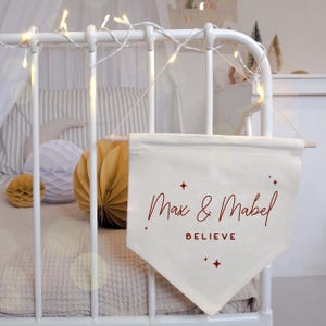 Könnte beinhalten: Cremefarbenes Banner mit den Namen "Max & Mabel" in roter Schrift, dem Wort "BELIEVE" und Sternakzenten. Das Banner hängt an einer Holzstange und ist an einem weißen Kinderbett befestigt. Dekorative Lichter sind darüber angebracht.