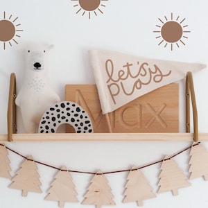 May include: A wooden shelf with a white polar bear stuffed animal, a wooden toy with black and white spots, a wooden sign with the word "Max" carved into it, and a white flag with the words "let's play" written in brown. The shelf is decorated with a garland of wooden Christmas trees and brown sun stickers on the wall behind the shelf.