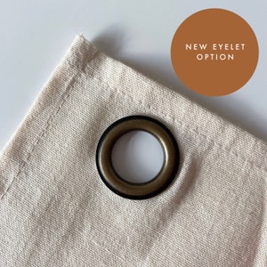Puede incluir: Primer plano de una esquina de tela beige con una arandela de bronce oscuro. La arandela es circular con un centro oscuro y un anillo exterior de bronce. Un c&iacute;rculo marr&oacute;n con el texto "NEW EYELET OPTION" est&aacute; en la esquina superior derecha.