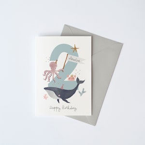 May include: A blue and white birthday card featuring a whale, octopus and crab. The number 2 is in the background with the name Amelia on a banner. The text "Happy Birthday" is at the bottom of the card.