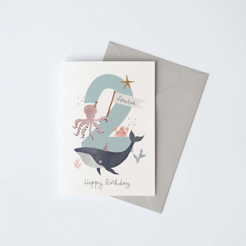 May include: A blue and white birthday card featuring a whale, octopus and crab. The number 2 is in the background with the name Amelia on a banner. The text "Happy Birthday" is at the bottom of the card.