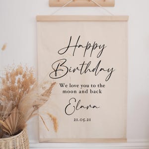 May include: A beige canvas wall hanging with the words "Happy Birthday" in elegant black script. Below, it reads "We love you to the moon and back" and "Elana" with the date 21.05.21. The canvas is held by wooden rods.