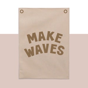 Puede incluir: Un tapiz de pared de lona beige con dos ojales en la parte superior. El tapiz tiene el texto "MAKE WAVES" impreso en marrón.
