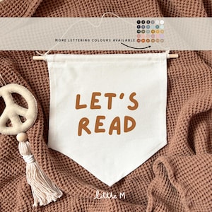 Peut inclure: Bannière en toile blanche avec le texte "Let's Read" en lettres brunes. La bannière est accrochée à un mur texturé brun.