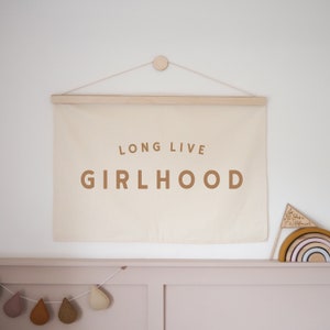 May include: A beige canvas banner with a wooden dowel at the top, hanging on a white wall. The banner has the text "LONG LIVE GIRLHOOD" printed in brown letters.