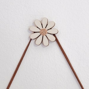May include: A white wooden daisy with a gold glitter centre hangs from two brown leather cords.
