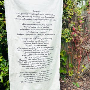 Op de afbeelding: Een witte stoffen banner met de tekst van Psalm 139 in zwarte letters. De banner hangt buiten, met groene bladeren en een hek op de achtergrond. De tekst is een religieuze passage.