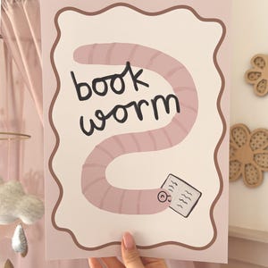 Pode incluir: Uma impressão rosa e branca com uma ilustração de desenho animado de um verme de livro. O verme é rosa e segura um livro, com as palavras "book worm" em preto. Possui uma borda marrom ondulada.