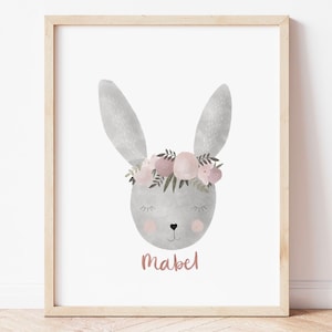 May include: A framed print featuring a watercolor illustration of a gray bunny with a floral crown. The bunny's face has closed eyes and rosy cheeks. The name "Mabel" is written in a cursive font below the bunny's face. The print is in a light wood frame.