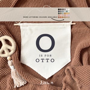Op de afbeelding: Een witte canvasbanner met een zwarte letter O en de tekst "O is for OTTO" in zwart. De banner hangt aan een bruin, getextureerd oppervlak. De tekst "little M" staat in de rechterbenedenhoek.