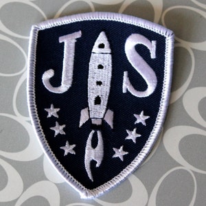 May include: A white embroidered patch with a navy blue background. The patch is shaped like a shield and features the letters "JS" in white with a white rocket ship in between the letters. The rocket ship has a white flame and there are white stars around the rocket ship.