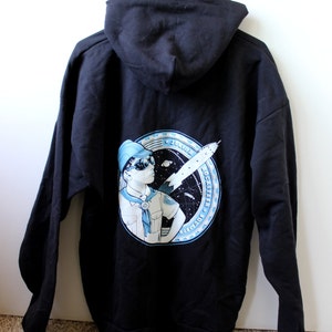 Puede incluir: Sudadera con capucha negra con un gráfico azul y blanco de una persona joven con uniforme de explorador y sosteniendo un cohete. El gráfico está rodeado por un círculo con el texto "Junior Rocketry Squad" en una fuente azul y blanca.