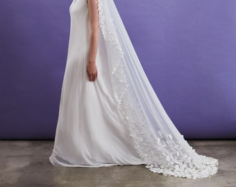 May include: A white wedding dress with a long, flowing skirt and a simple, sleeveless bodice. The bride is wearing a white veil with delicate floral embellishments.