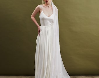 May include: A woman wearing a white satin wedding dress with a long, flowing veil. The dress has a simple, elegant design with a V-neckline and a flowing skirt. The woman is standing in front of a green background.