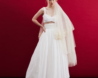 May include: A woman wearing a white wedding dress with a lace trim veil. The dress has a fitted bodice and a flowing skirt. The woman is standing in front of a red background.