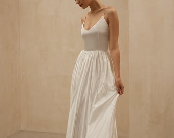 May include: A simple white wedding dress with a fitted bodice and a flowing skirt. The dress has a V-neckline and thin straps.