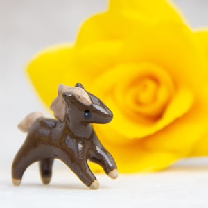 Op de afbeelding: Een klein bruin keramiek paardje met een lichtbruine manen en staart. Het beeldje staat op een wit oppervlak met een gele roos op de achtergrond.