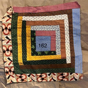 May include: A square patchwork quilt with a central design featuring the number "162". The quilt incorporates various fabrics in shades of red, blue, yellow, green, and brown. The edges are finished with patterned borders.