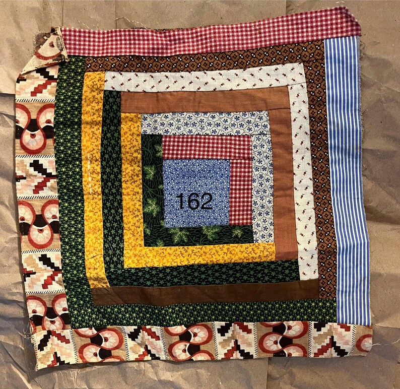 May include: A square patchwork quilt with a central design featuring the number "162". The quilt incorporates various fabrics in shades of red, blue, yellow, green, and brown. The edges are finished with patterned borders.