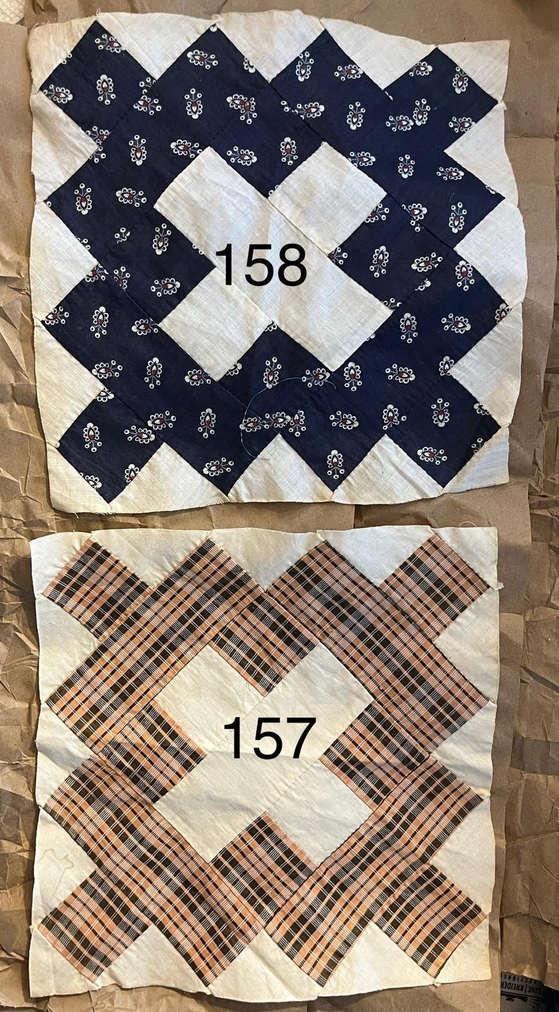 May include: Two vintage quilt blocks. One block features a navy blue floral pattern with the number "158" in the center. The other block has a plaid pattern in shades of orange, black, and white, with the number "157".