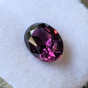 May include: An oval-shaped, faceted gemstone with a deep purple hue, transitioning to vibrant pink in the center. The gem is set against a white, textured background, showcasing its polished surfaces and internal reflections.