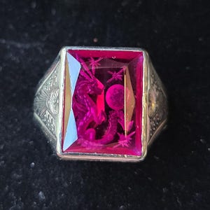 May include: A silver ring with a large, rectangular, ruby-colored gemstone. The gemstone is intricately carved with a dragon, stars, and a moon. The ring's band has detailed patterns.