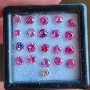 Puede incluir: Una colección de gemas redondas y facetadas en varios tonos de rosa y púrpura, exhibidas en una caja cuadrada negra con interior blanco. Las gemas son de diferentes tamaños y tonalidades, creando una disposición visualmente atractiva.